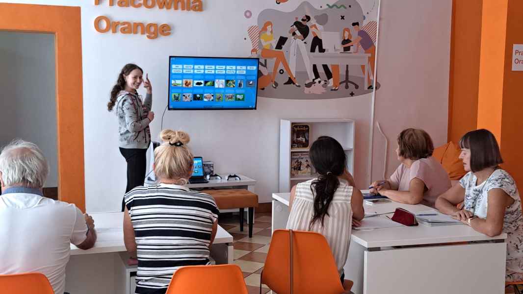 zdjęcie bibliotekarki pokazującej grupie seniorów zadanie na ekranie telewizora