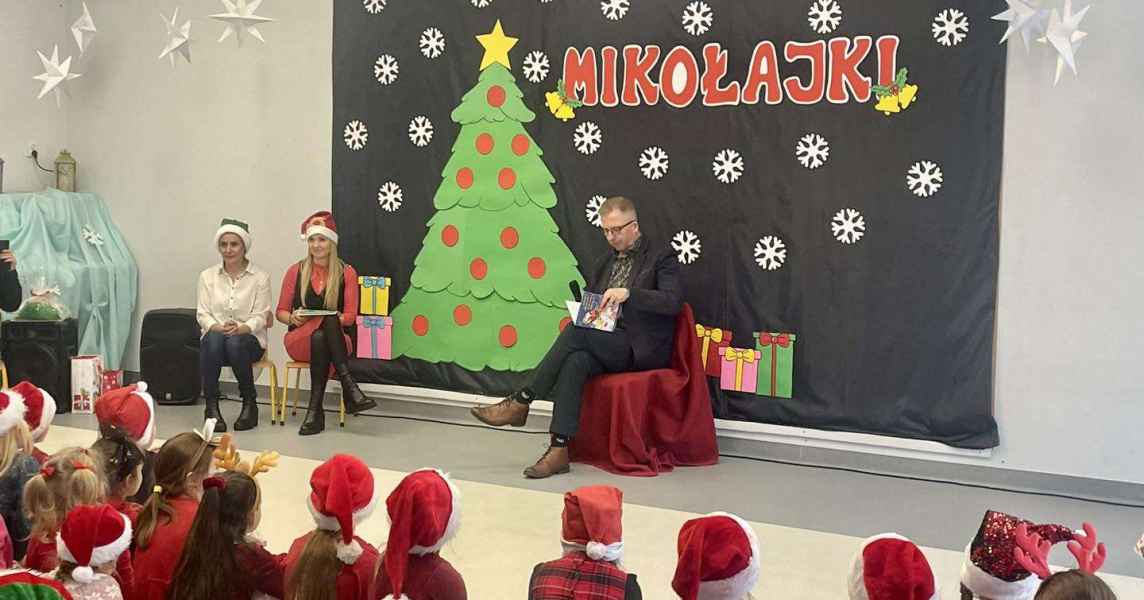 zdjęcie burmistrza czytającego dzieciom bajki,  z boku bibliotekarka i dyrektor biblioteki , przed nimi dzieci 