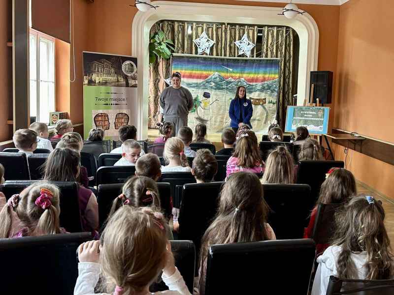 zdjęcie dzieci na widowni słuchających bibliotekarki i aktora na scenie