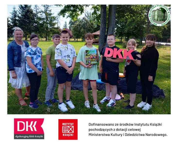 zdjęcie uczestników klubu DKK trzymających napis DKK i książkę z wychowawczynią na trawie, pod spodem belka logotypowa DKK, Instytutu kultury i napis dofinansowano ze środków Instytutu Książki, po prawej logo biblioteki