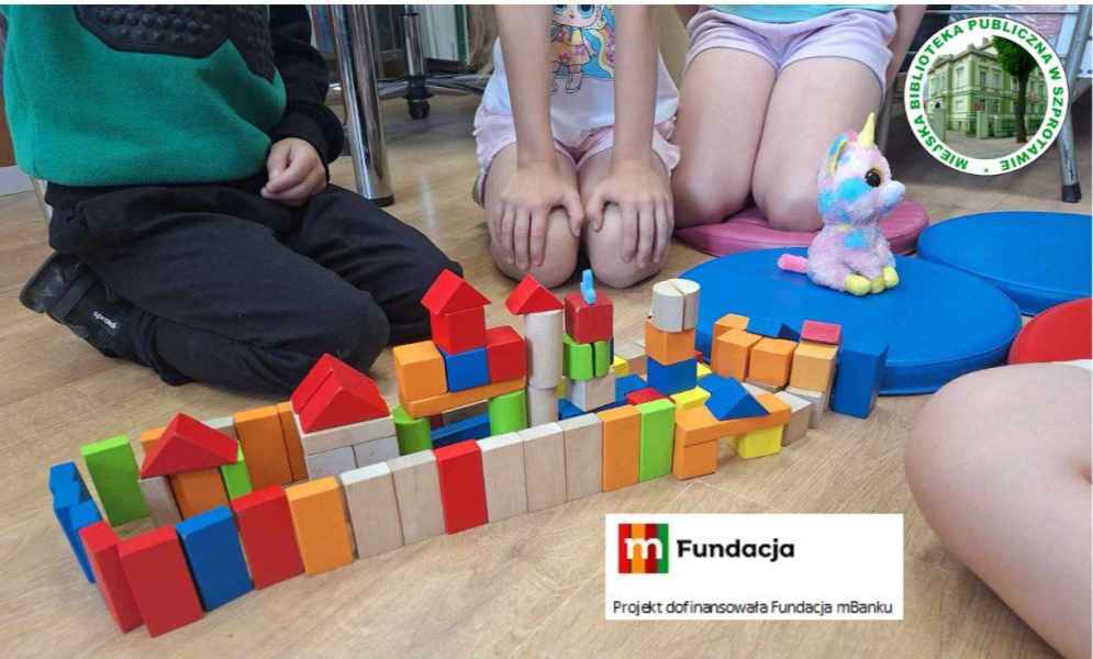 zdjęcie ułożonej budowli z klocków na podłodze, w tle dzieci, na górze logo biblioteki, na dole logo mfundacji i napis Projekt dofinansowała fundacja mbanku