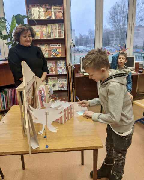 zdjęcie chłopca piszącego na kartce życzliwe słowo, obok bibliotekarka i inny uczeń