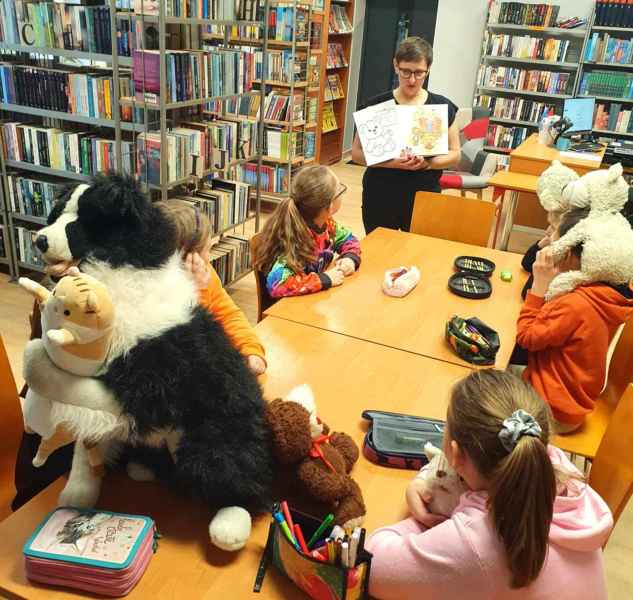 zdjęcie bibliotekarki pokazującej kolorowanki dzieciom siedzącym przy stole z pluszakami