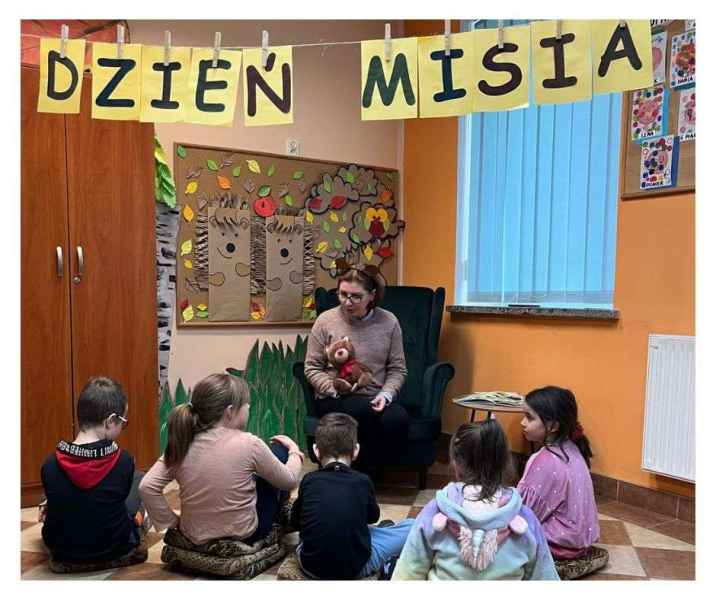 zdjęcie bibliotekarki w fotelu pokazującej dzieciom pluszowego misia, nad nimi wisi napis dzień misia