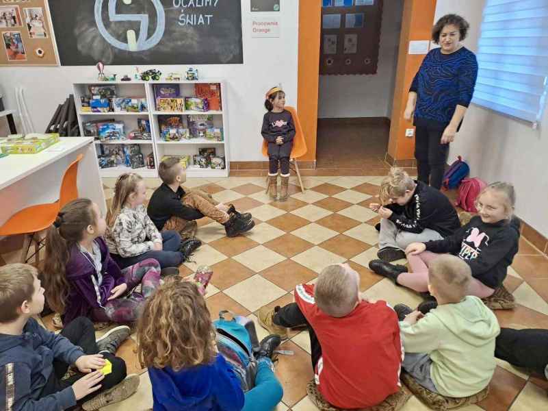 zdjęcie dzieci siedzących na podłodze i dziewczynki na krześle, z boku bibliotekarka tłumacząca zasady zabawy