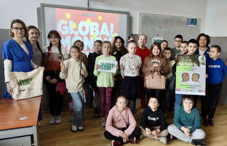 grupowe zdjęcie bibliotekarki, nauczycielki i dzieci z gadżetami projektu global money week i plakatem biblioteki
