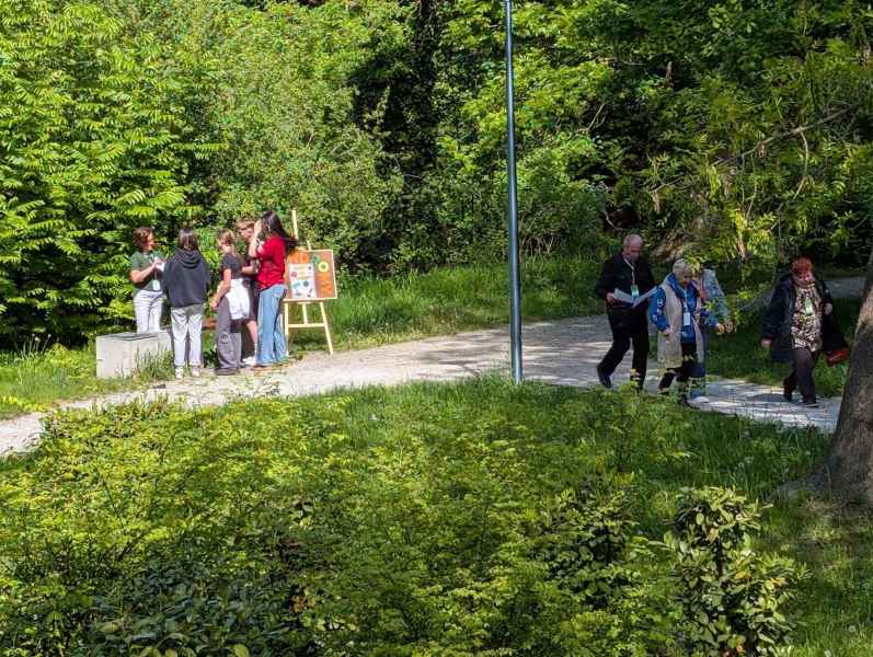 zdjęcie grup uczestników gry miejskiej podczas opuszczania stanowiska i słuchania bibliotekarki w parku