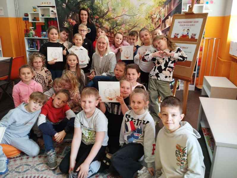 grupowe zdjęcie dzieci z autorką Mają Gnach i bibliotekarką na tle bajkowego baneru z książkami