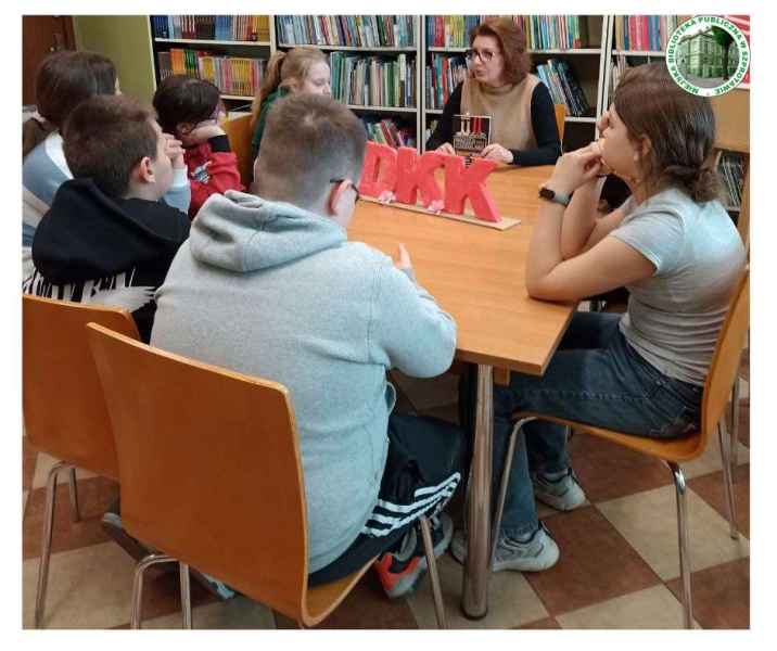zdjęcie młodzieży i bibliotekarki z książką przy stoliku, na nim napis DKK, po prawej logo biblioteki