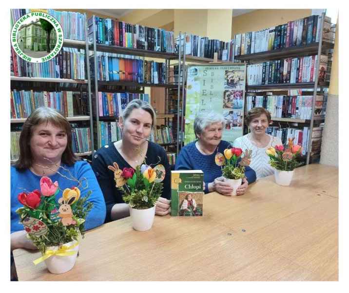 zdjęcie czyteniczek przy stoliku z własnoręcznie wykonanymi stroikami i książką Chłopi reymonta, po lewej logo biblioteki