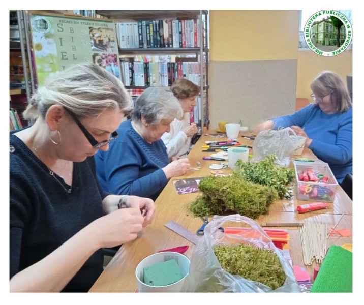 zdjęcie czytelniczek przy stoliku podczas przyklejania mchu chrobotka do gąbki florystycznej, po prawej logo biblioteki