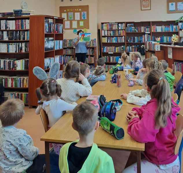 zdjęcie bibliotekarki pokazującej dzieciom przy stolikach ilustracje z teatrzyku kamishibai